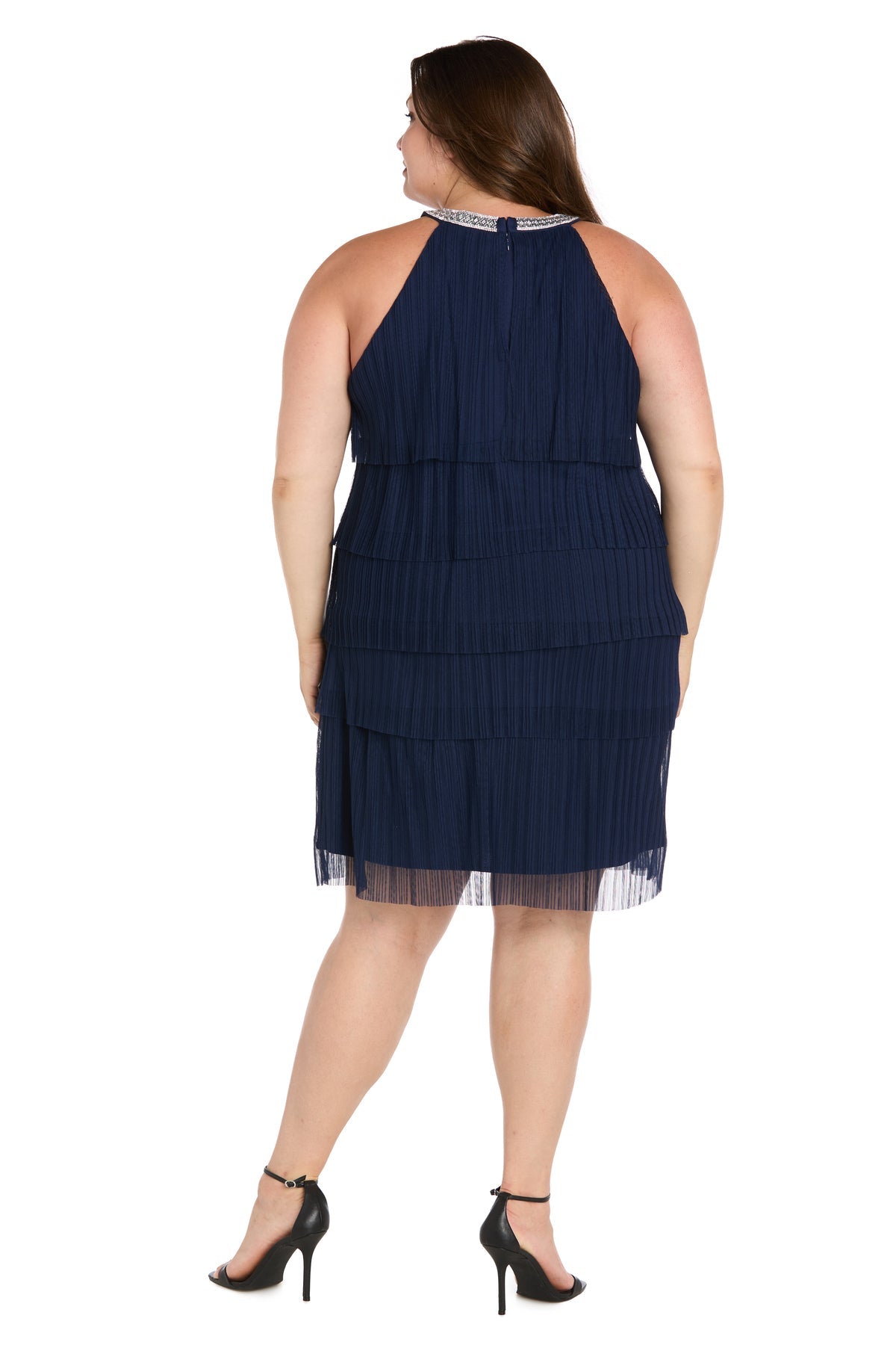 A woman wearing the R&M Richards 3744W plus-size navy party dress with tiers and black heels stands facing away against a white background.